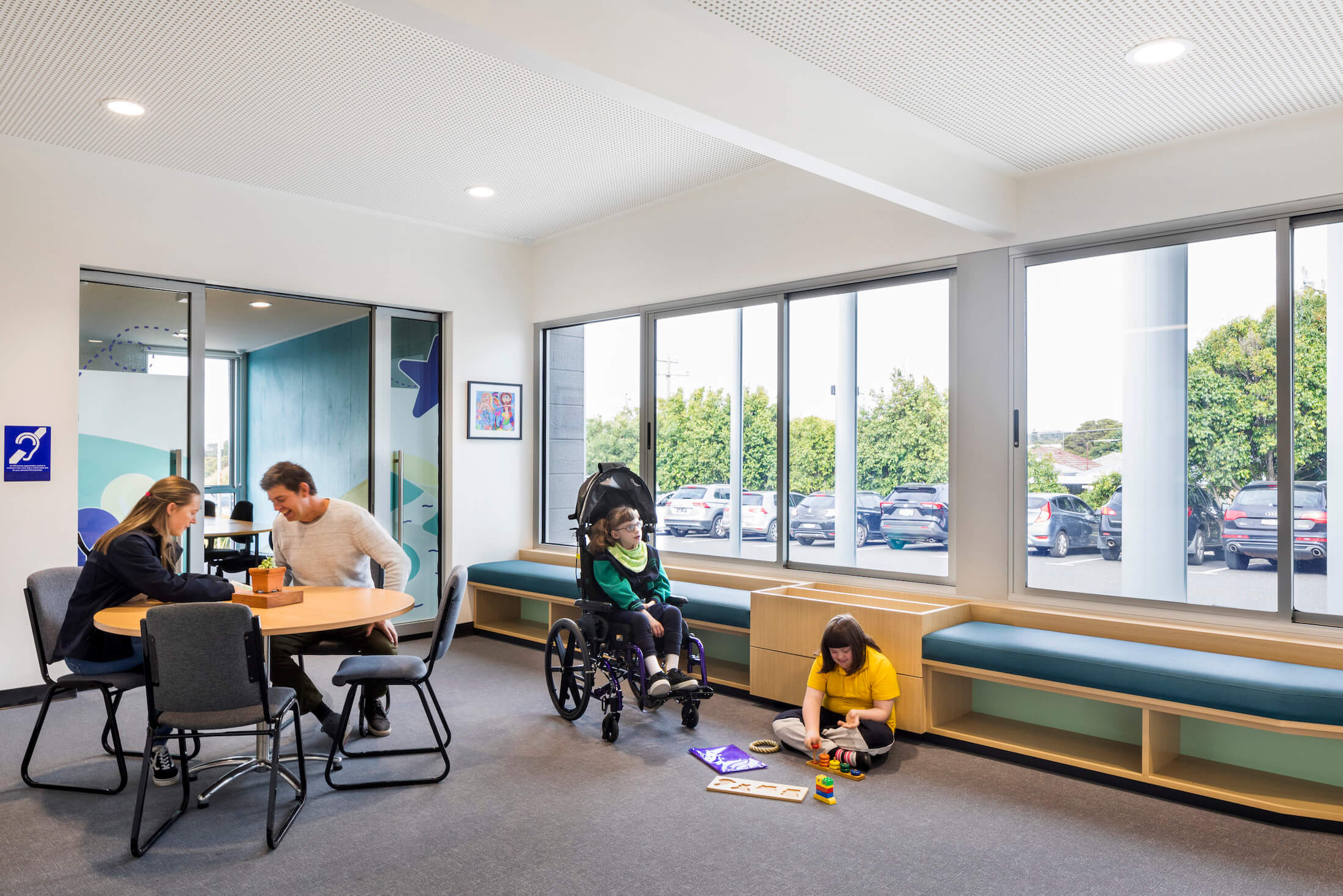 Adults at table in breakout area next to boardroom and reception, children sit in wheelchair and play with blocks on the floor
