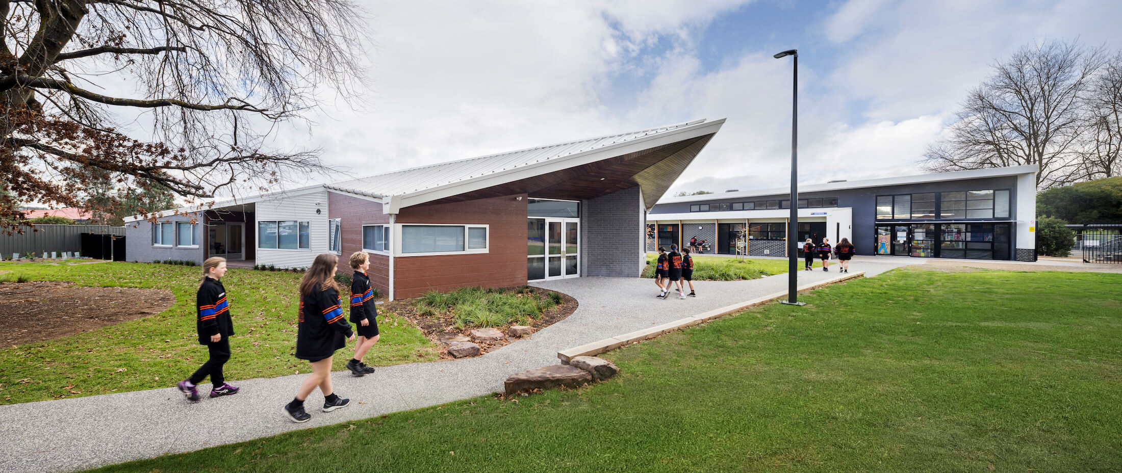 Panoramic image of students walking on path to new school building entry with pointed canopy and classrooms beyond