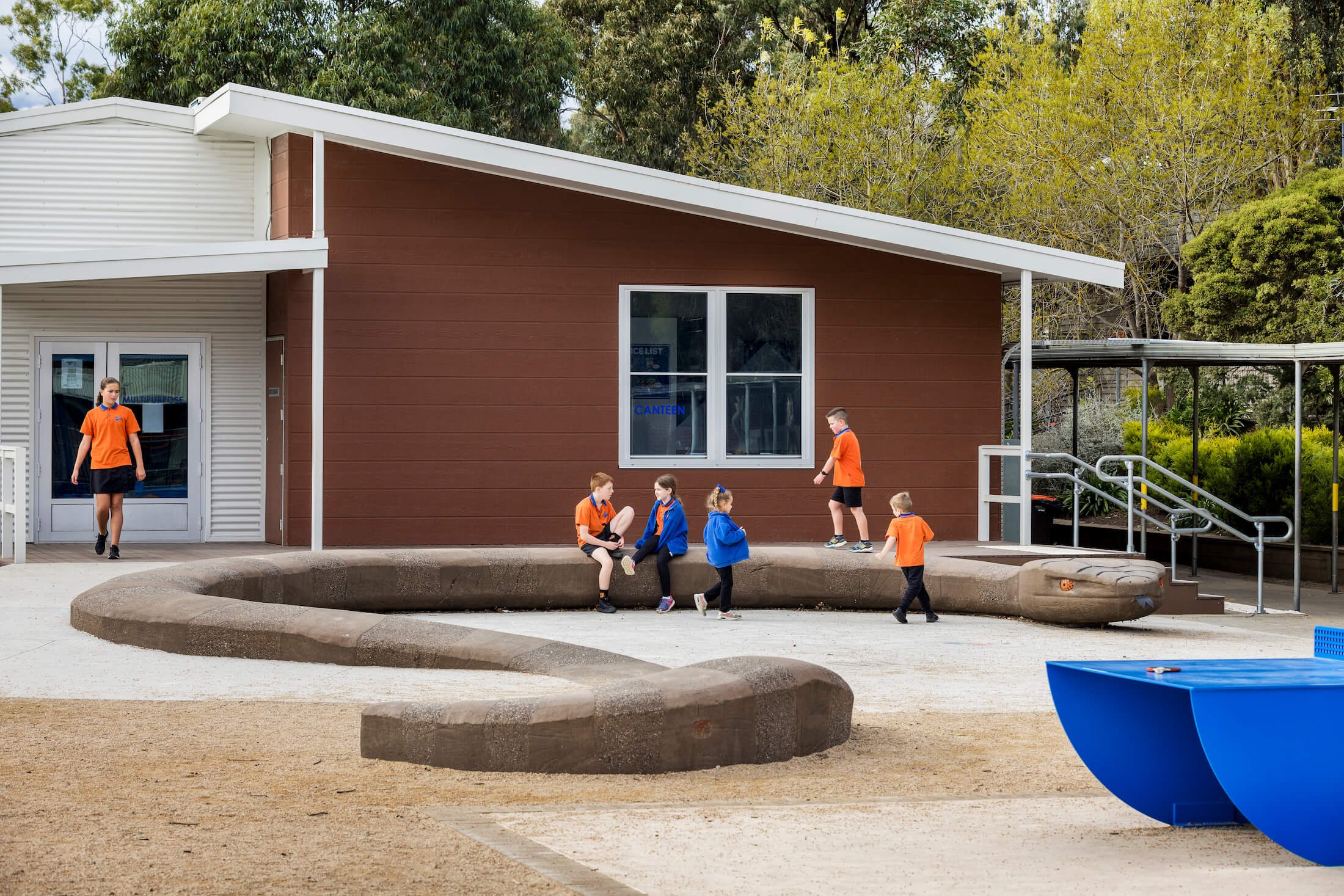 Students sit, walk and play on winding snake seat in playground, school building and established landscaping in the background