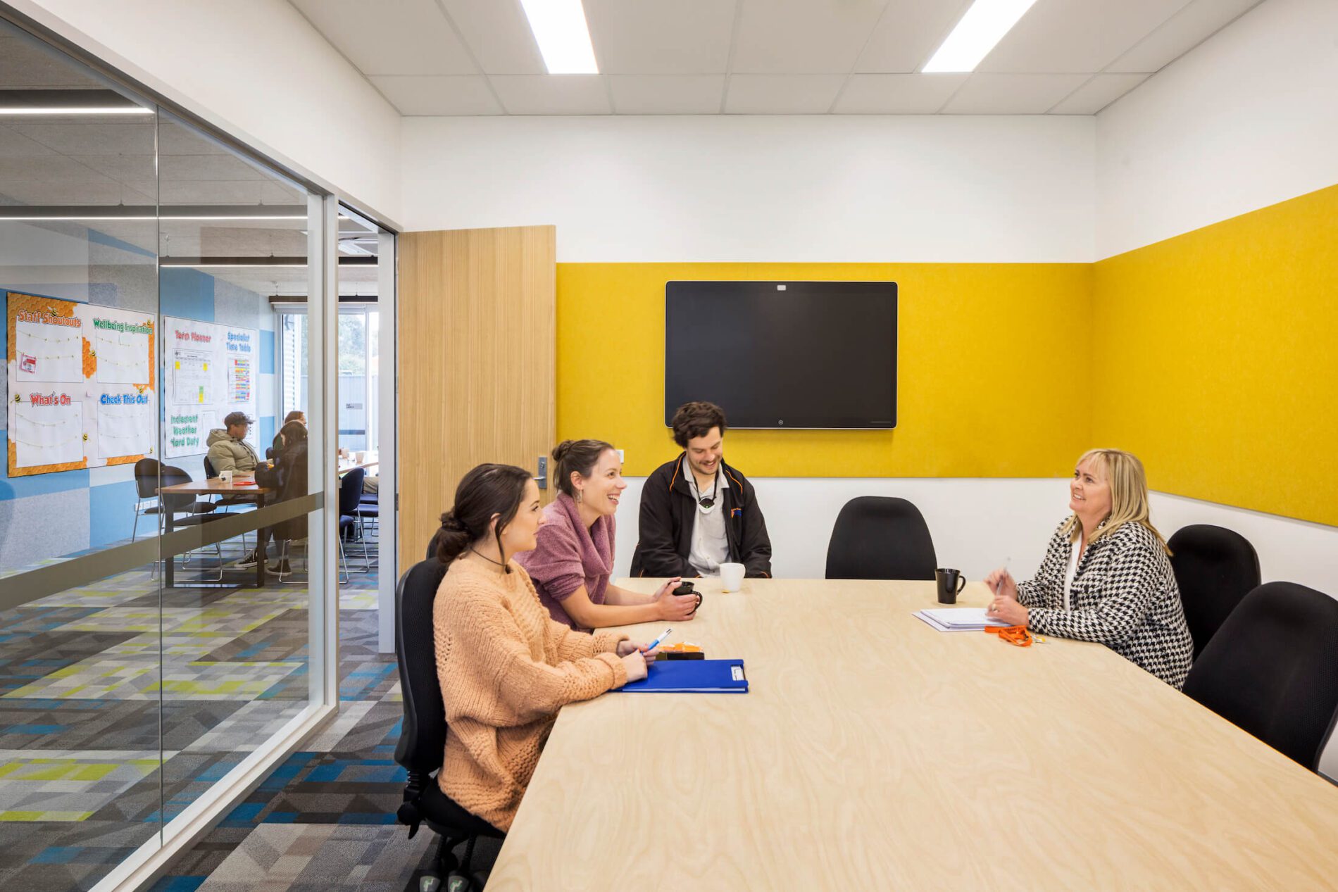 Four school staff sit at table in meeting room with yellow wall panel detail and television screen