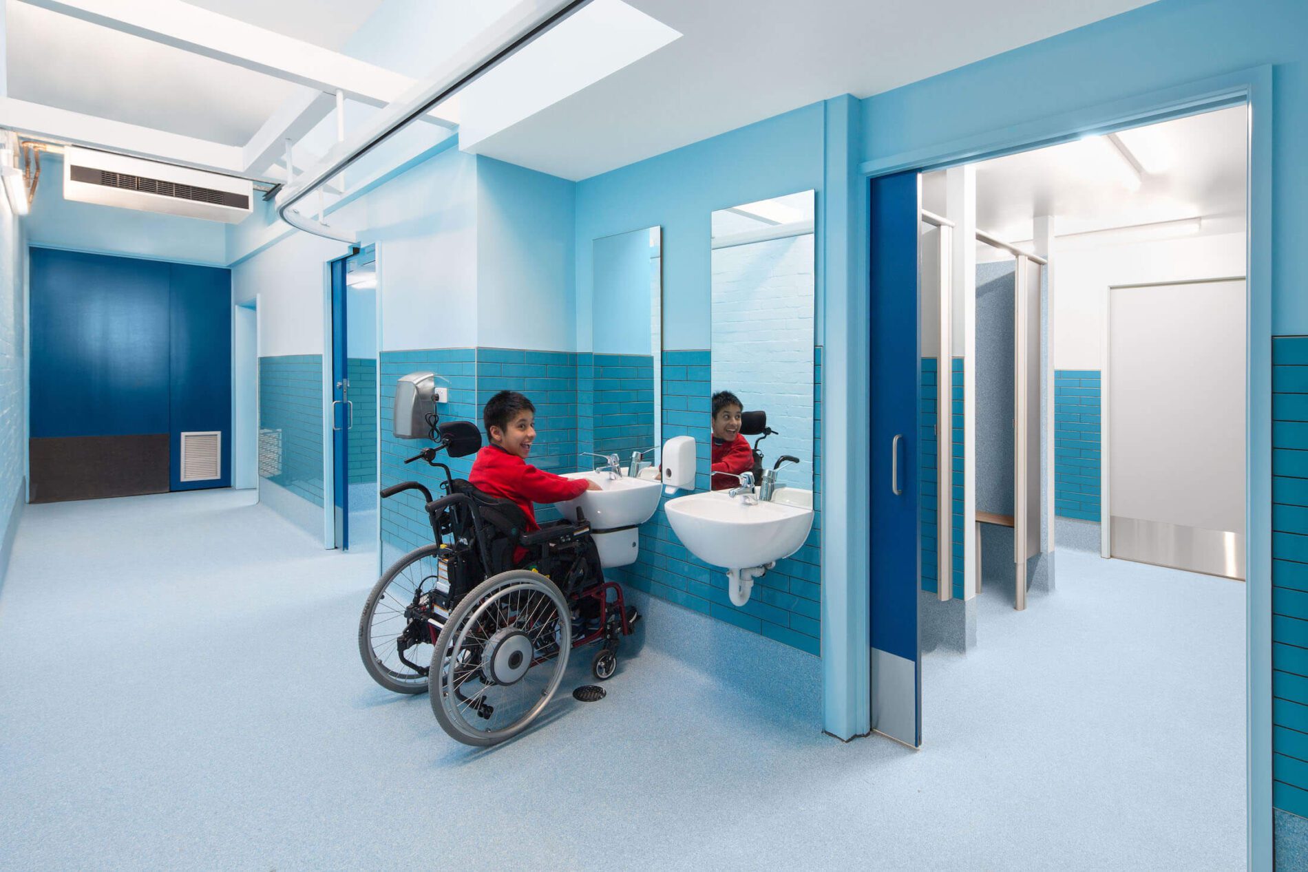 Student in wheelchair uses basin in blue toilet block, smiles at camera