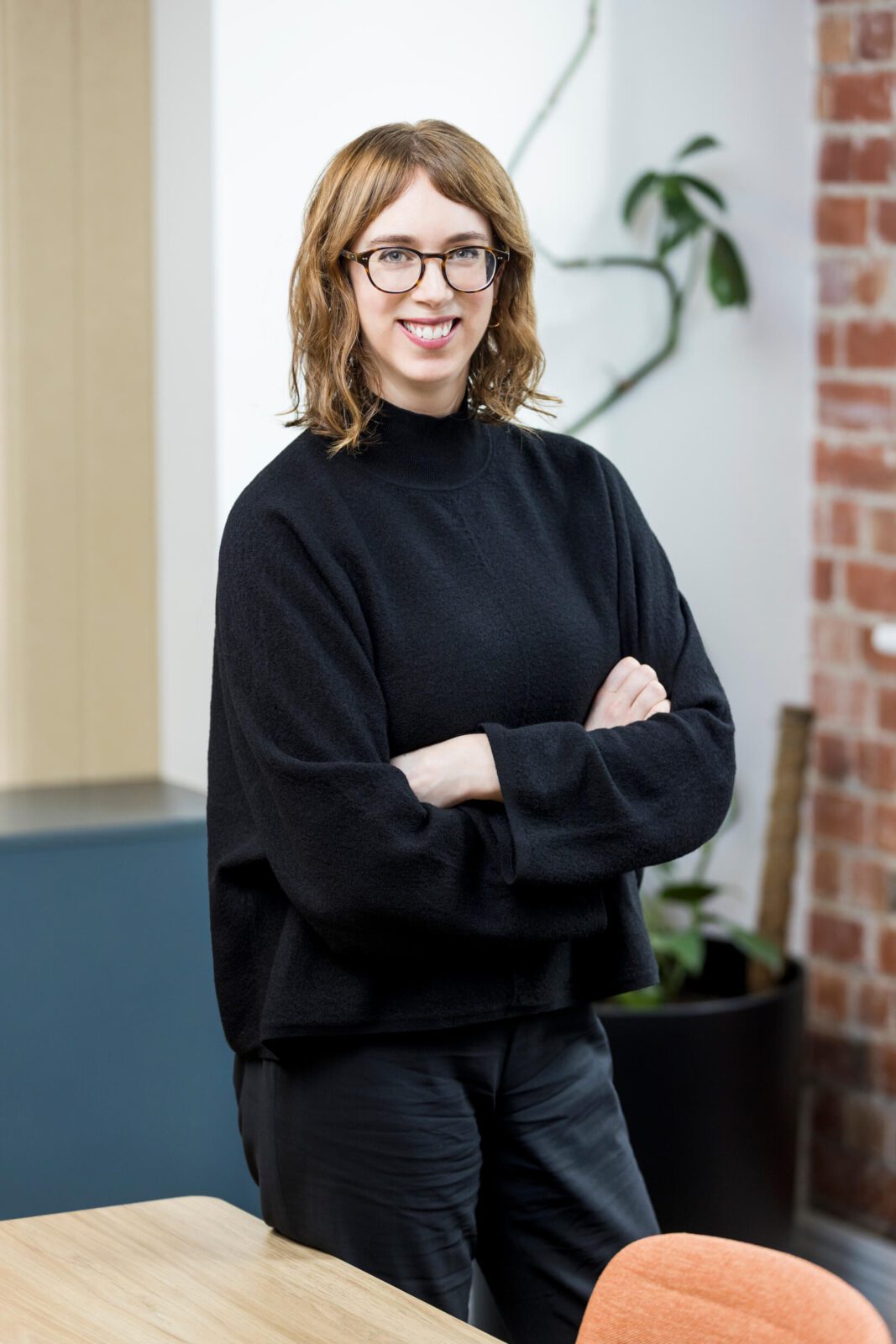 Woman in black high neck knit, tortoiseshell glasses, dark pants, stands cross-armed, smiling broadly to camera