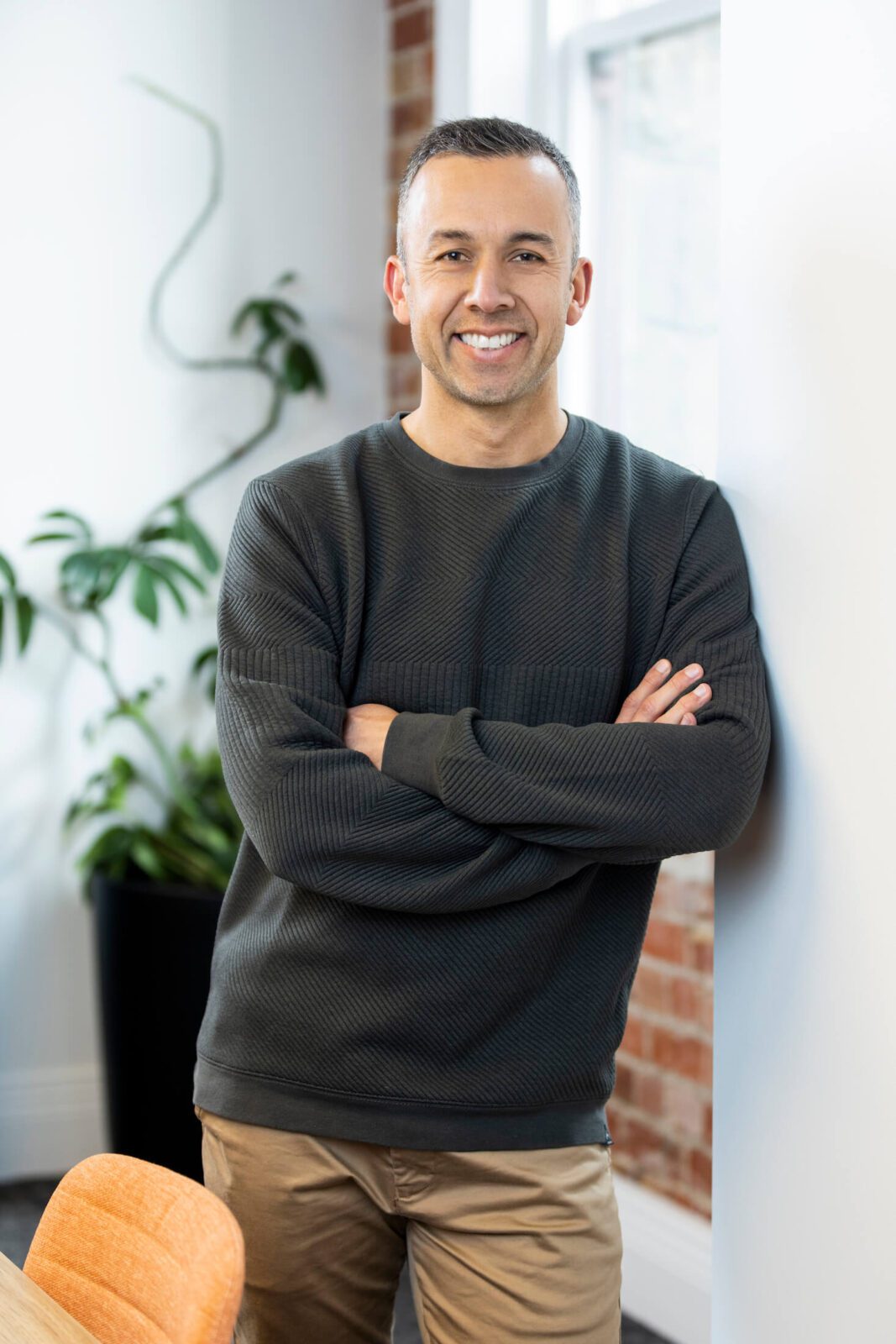 Man in grey knitted pullover and chinos leans against office wall, arms crossed, smiling broadly to camera