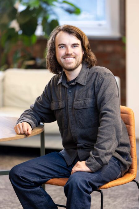 Man with shoulder length hair in grey long sleeve shirt and blues jeans seated on orange chair smiles broadly to camera