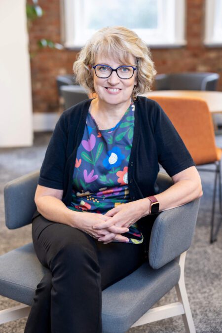 Woman in colourful top, black cardigan, wearing fine blue glasses and hoop earrings sits cross-legged in grey office armchair, smiling broadly to camera