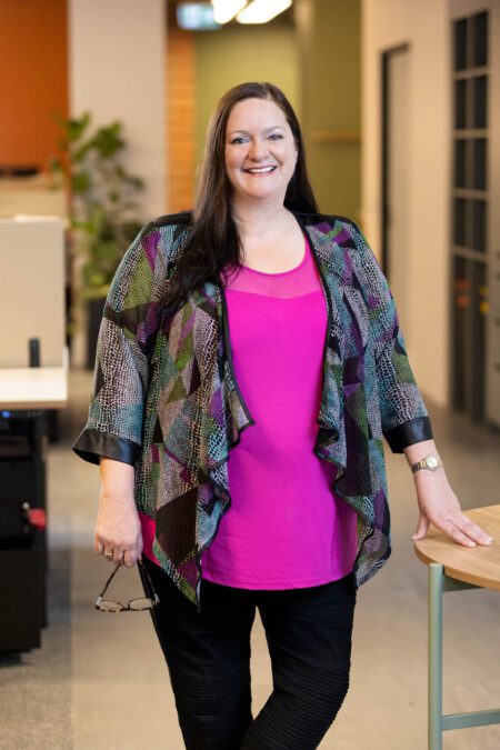 Woman with long dark hair in sheer, colourful jacket, bright pink top and black pants leans against office table, smiles broadly to camera