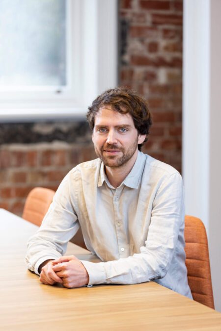 Man in natural shirt sits at boardroom table, smiles gently to camera