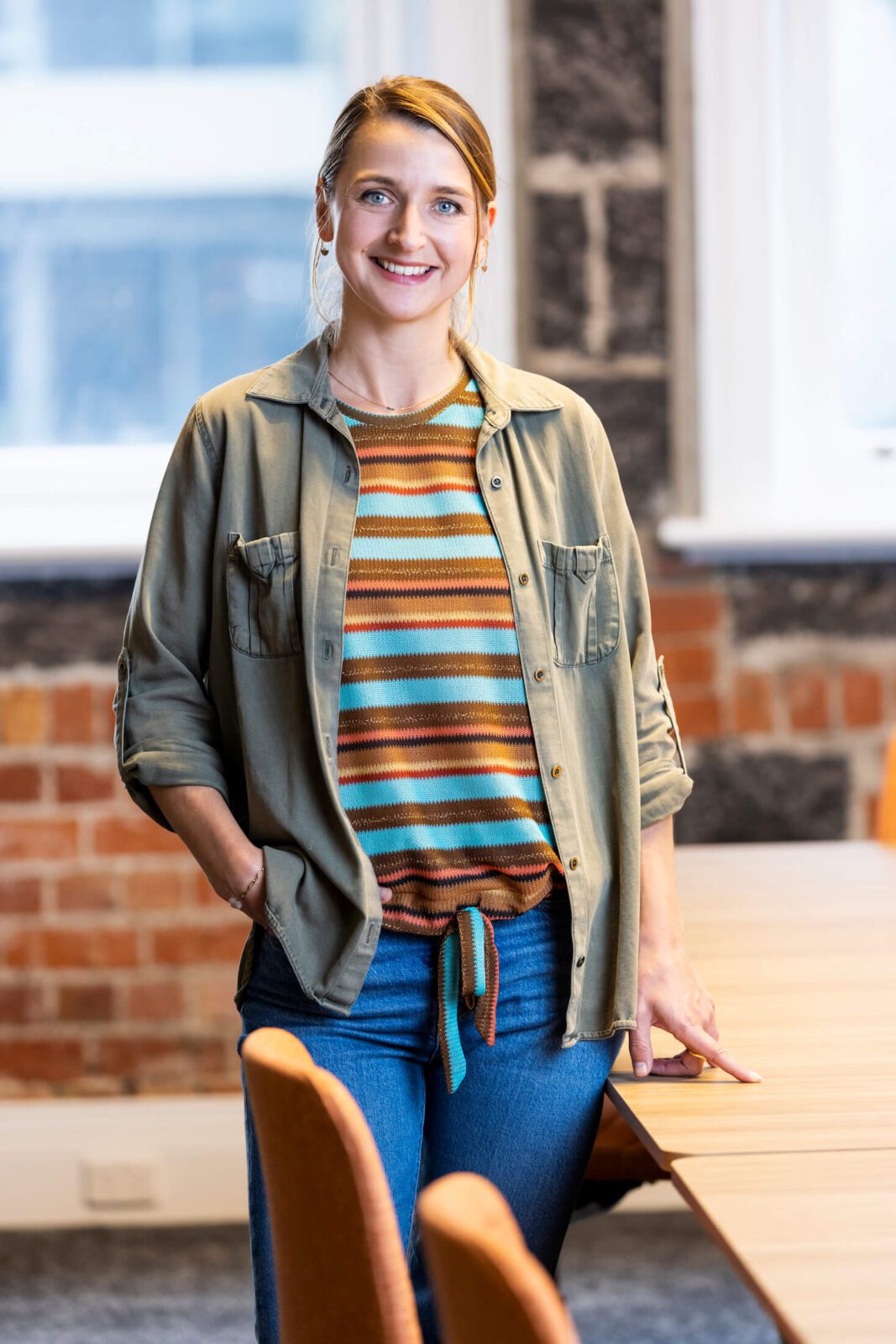 Woman in striped colourful top, army green shirt, blue jeans smiles broadly to camera leaning on boardroom table