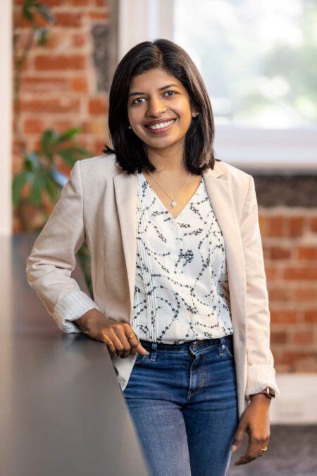 Woman with white v-neck shirt, cream blazer and blue jeans smiles broadly to camera