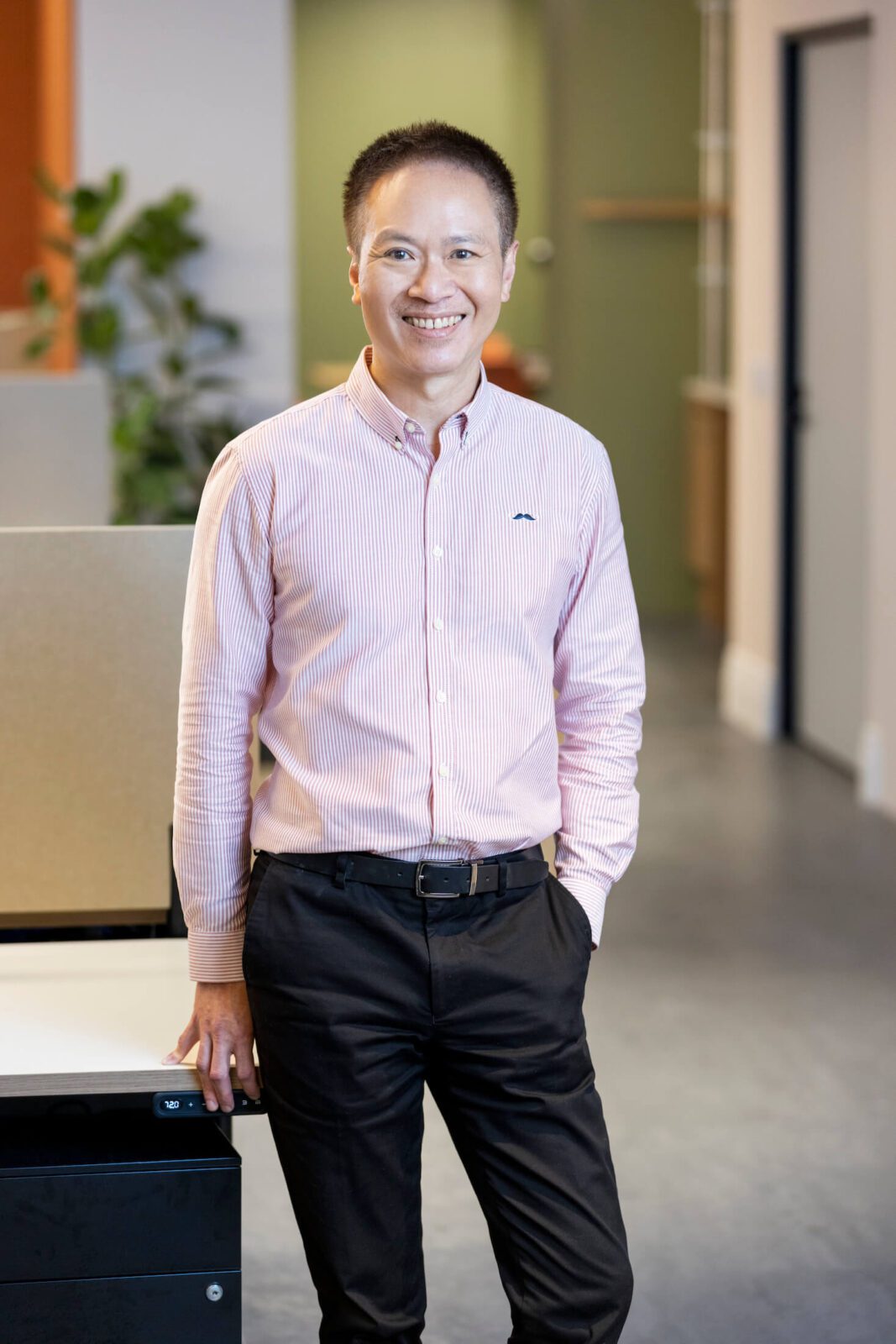 Man in pink pin-striped shirt, black pants, leans against desk in office, smiles broadly to camera