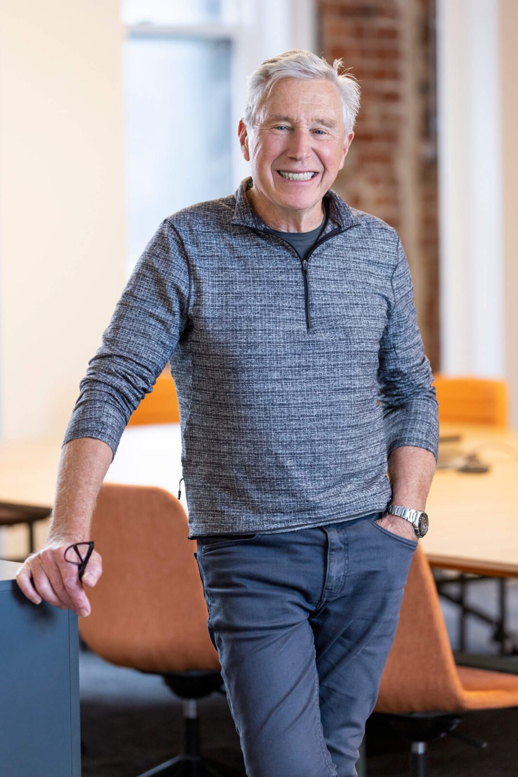 Man with grey half zip jumper, grey jeans, eyeglasses in hand, leans against in boardroom, smiles broadly to camera