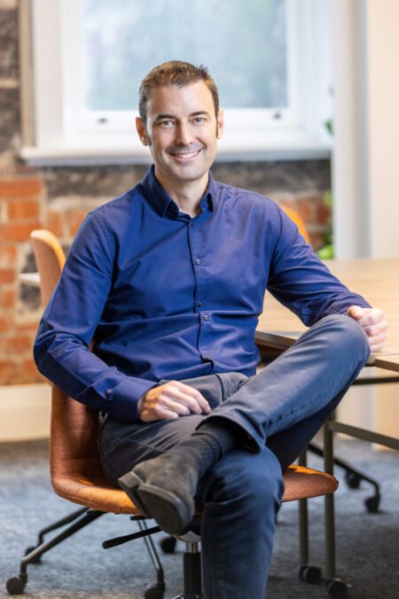 Man in blue shirt, dark pants and boots, sits cross-legged at boardroom table, smiles broadly to camera