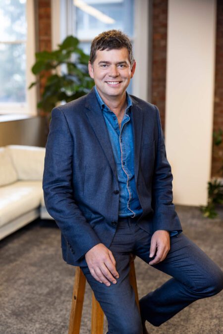 Man in blue chambray shirt, blue blazer and pants sits on stool, smiles broadly to camera