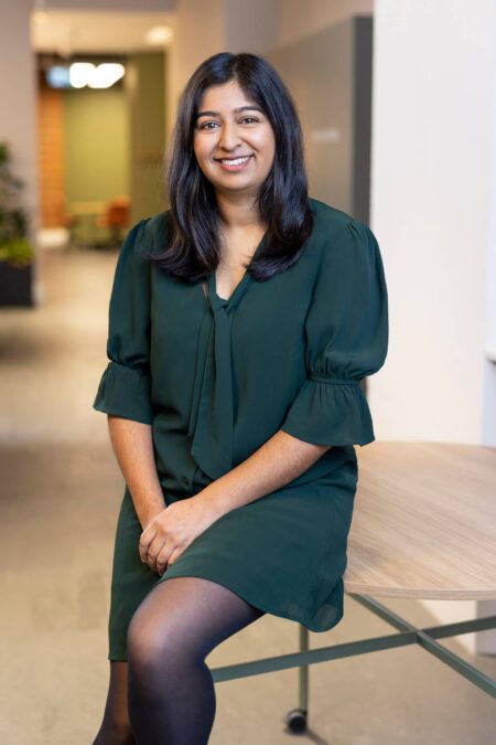 Woman with long dark hair, forest green dress, perches against office table, smiles to camera