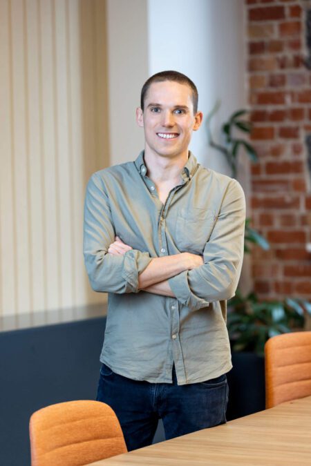 Man in light army green shirt, dark jeans stands arms crossed against boardroom table, smiles broadly to camera