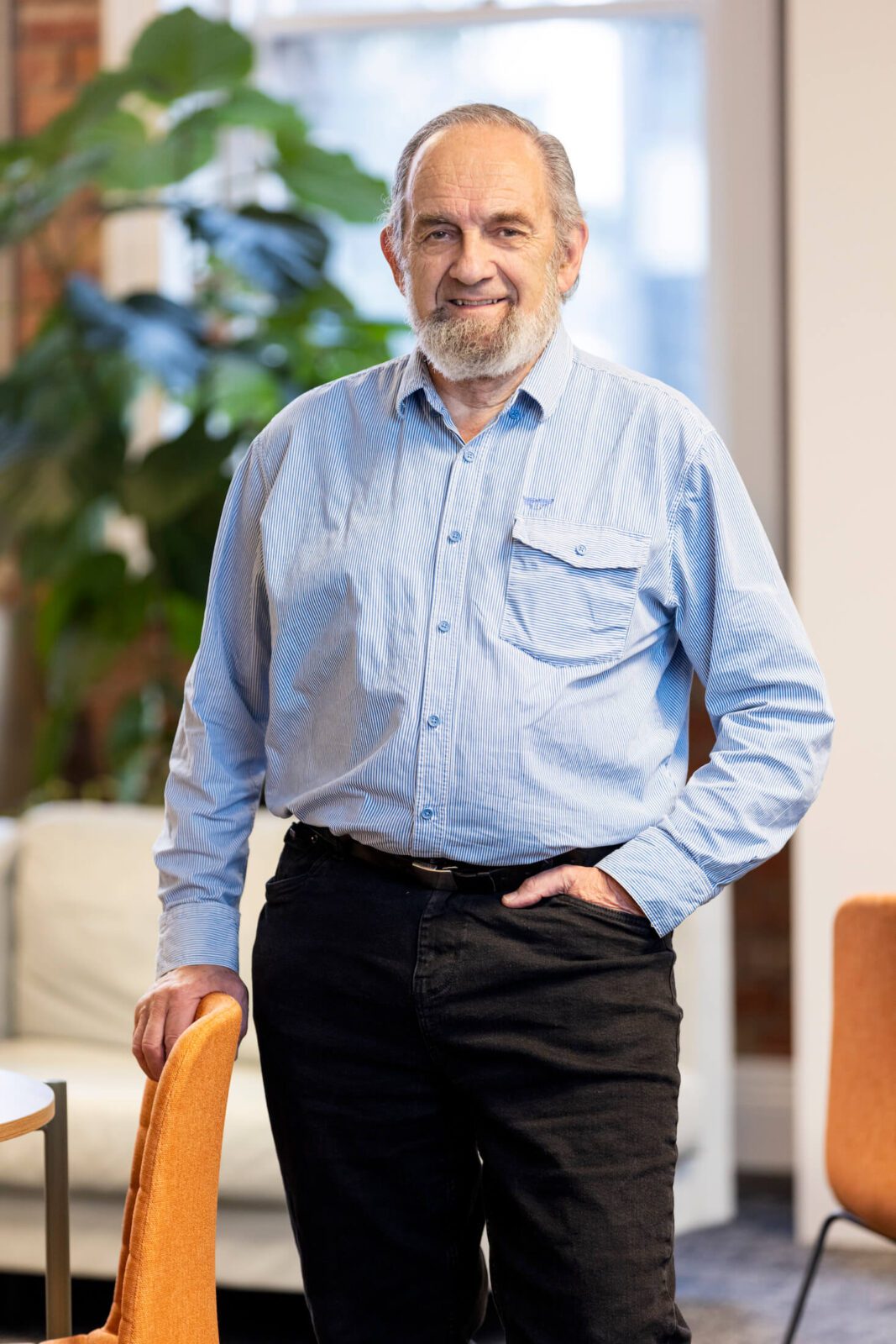 Man in light blue pinstriped shirt, dark jeans, with full grey beard and grey hair, leans against office chair, smiles at camera