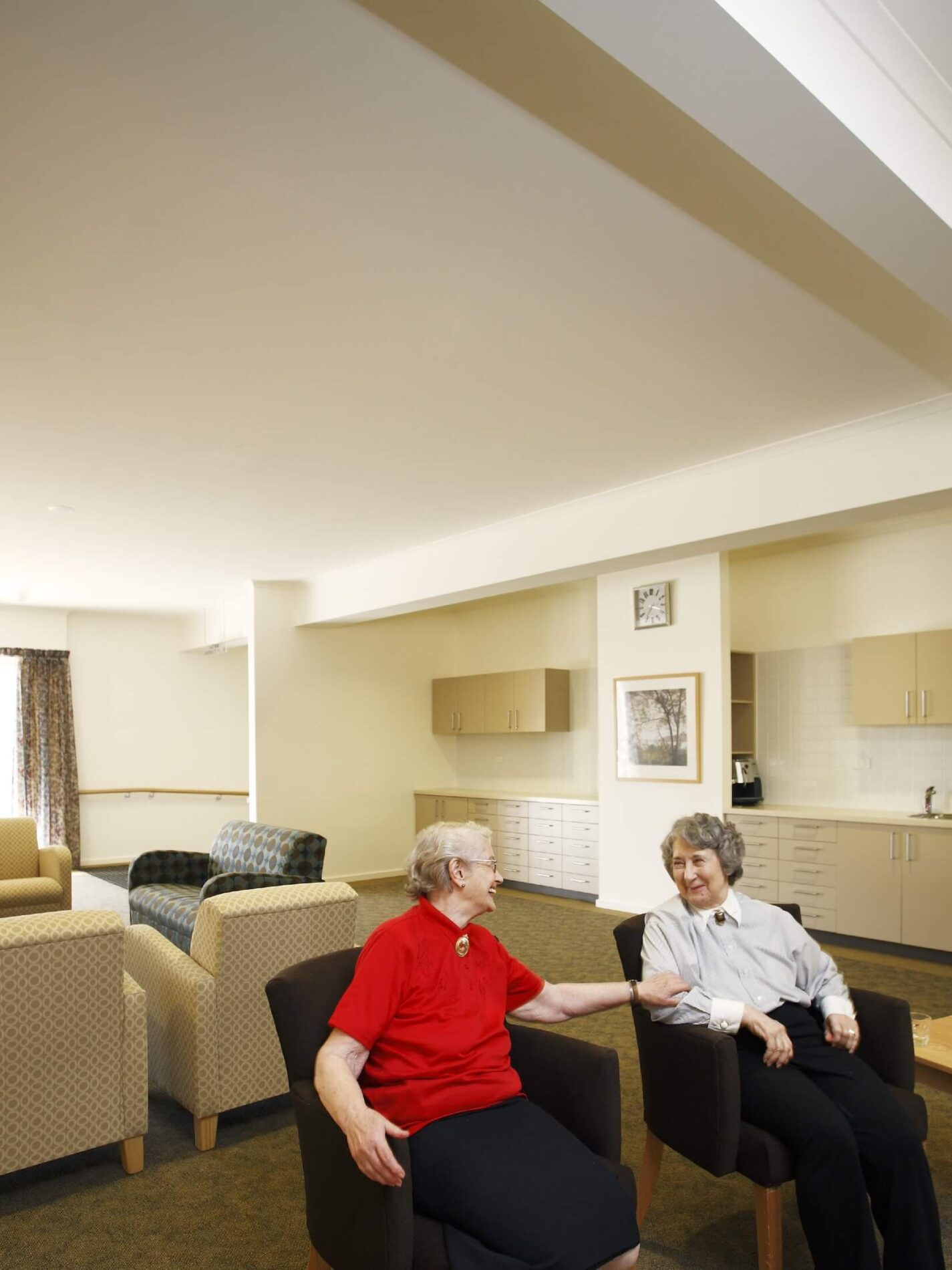 Two older women laugh and chat in armchairs in living space with kitchenette beyond