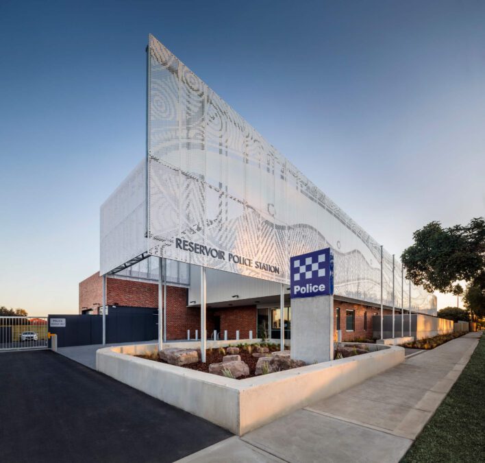 Late afternoon view of Reservoir Police Station with perforated steel screen facade displaying first nations artwork. Brick building with concrete planters.