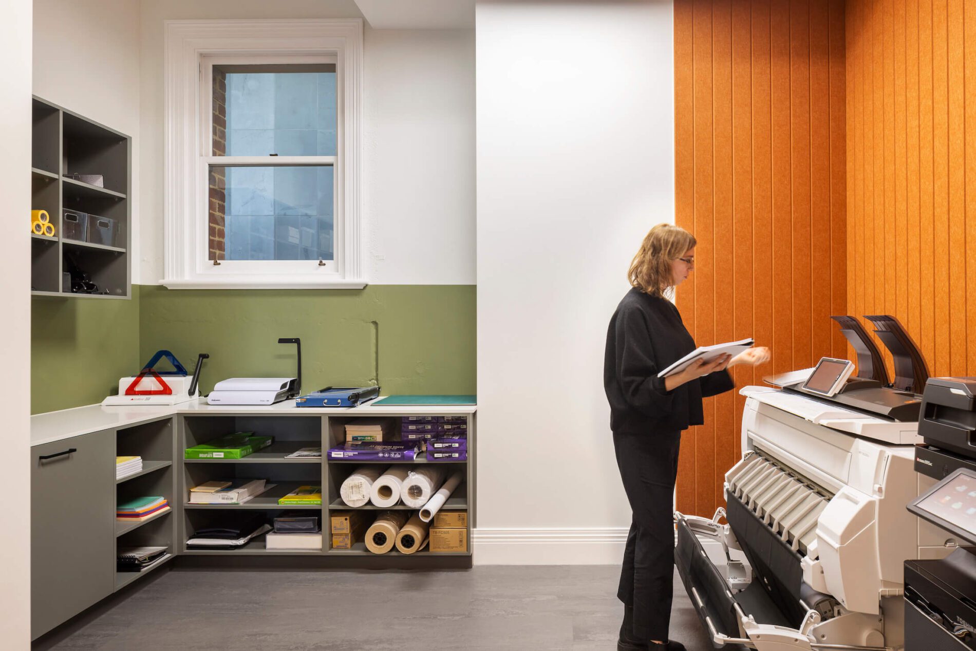Woman in black stands at printer in office utility room. Paper storage, guillotines in background. Orange vertical panelling and green feature walls