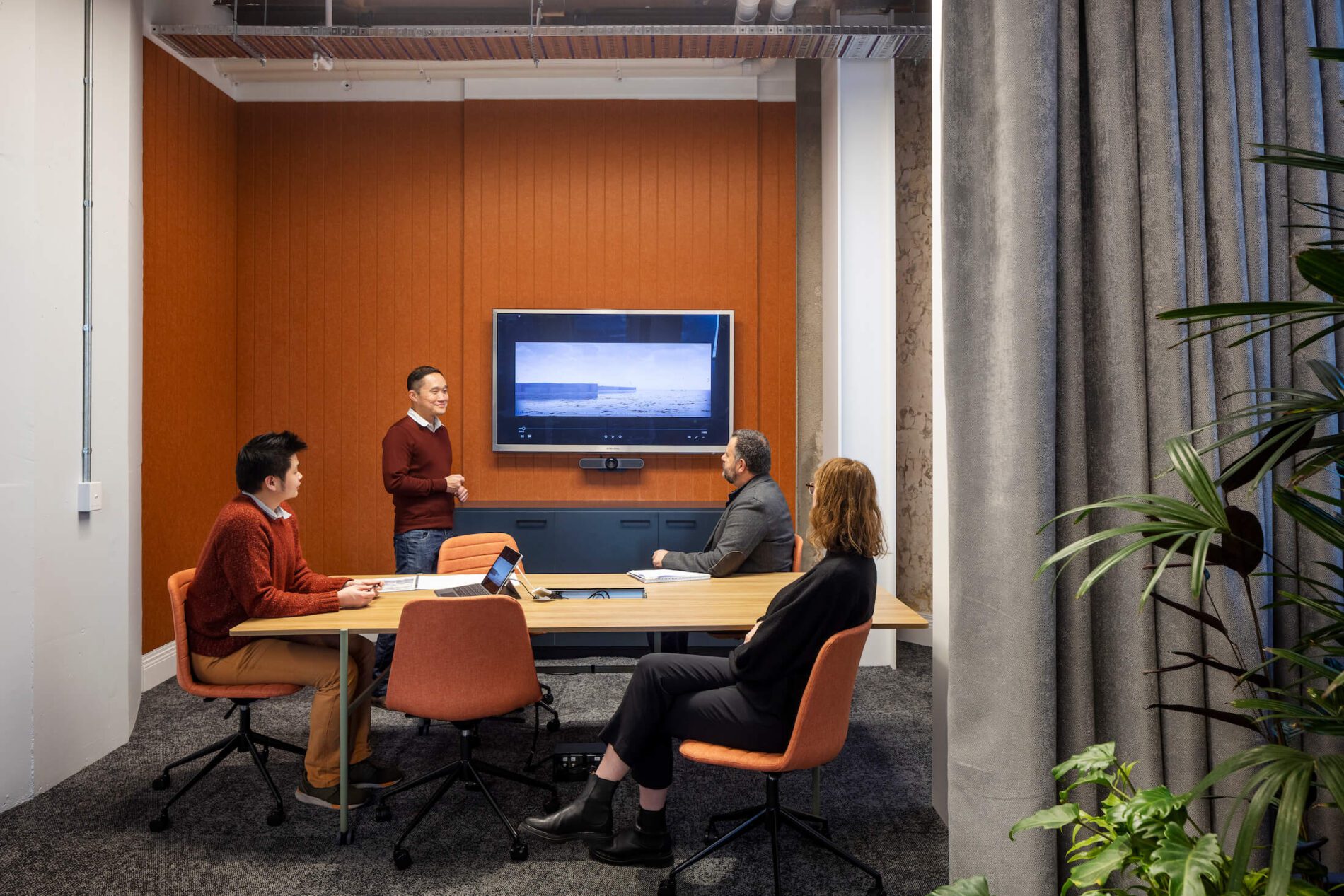 Three staff members in a meeting room sit at a table listening to another staff member present, using a video display.