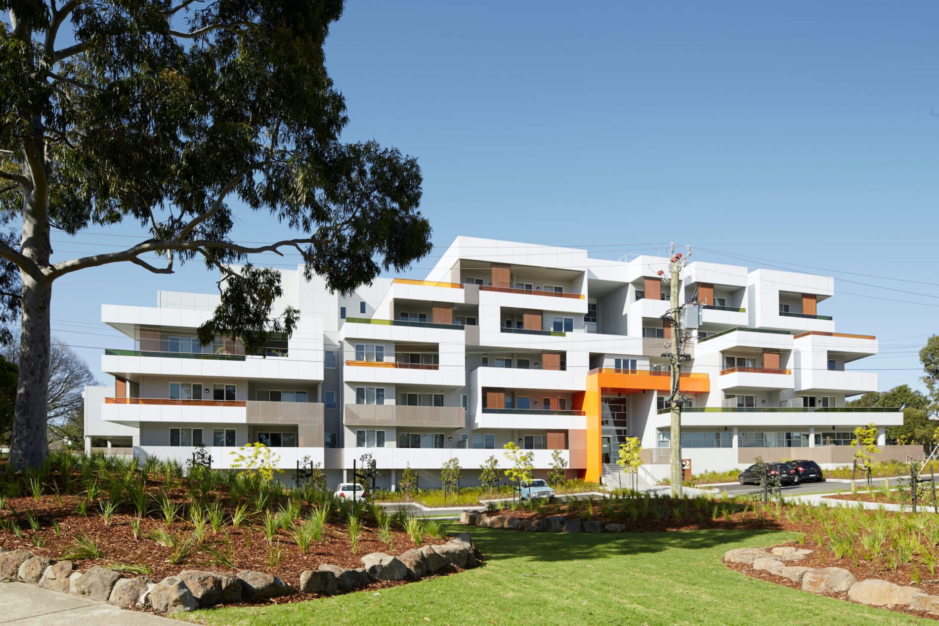 A white multi-storey apartment building with orange cladding feature elements, native landscaping and established gums out front