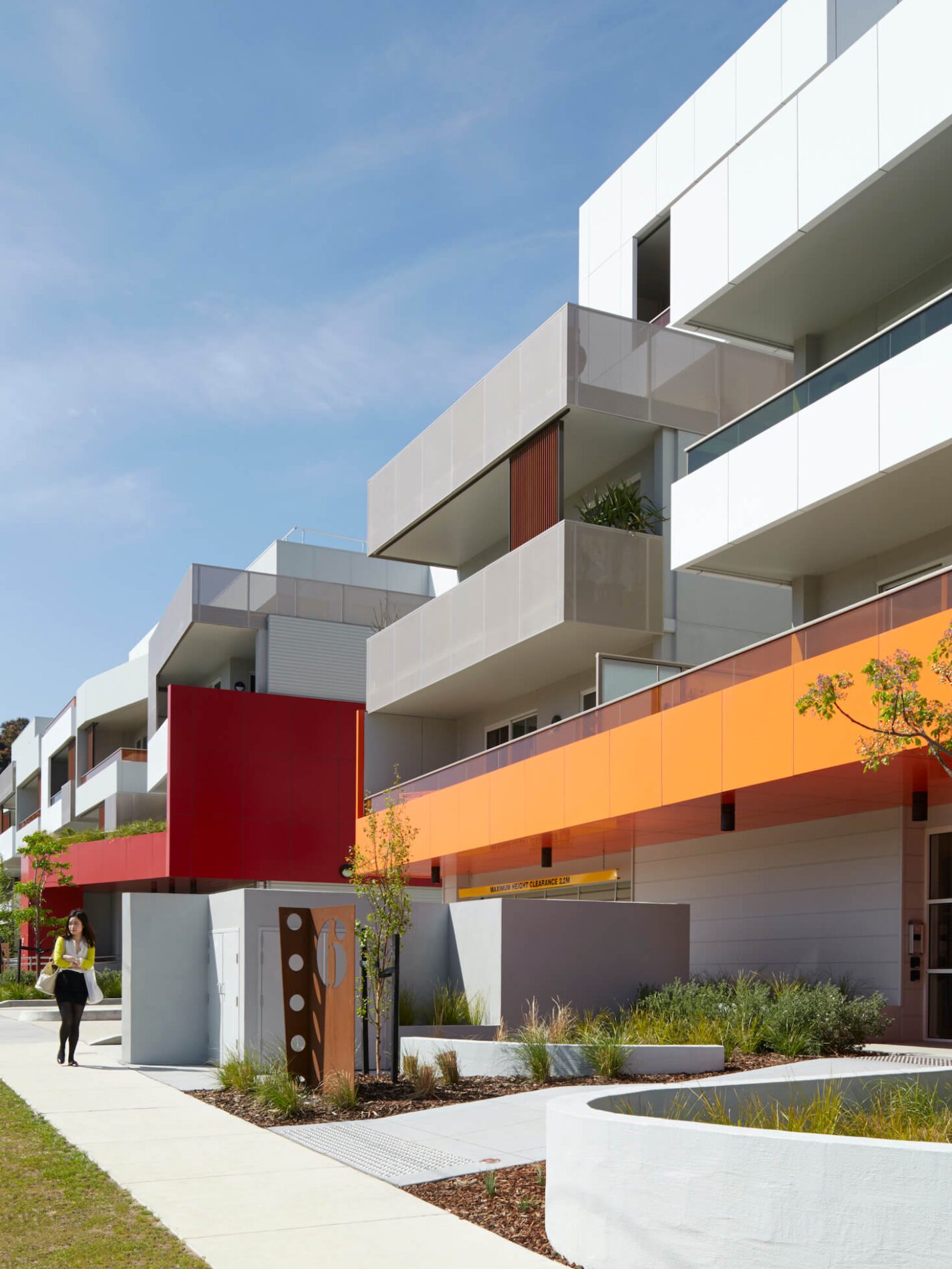 A white multi-storey apartment building with red and orange cladding feature elements, pedestrians and native landscaping out front