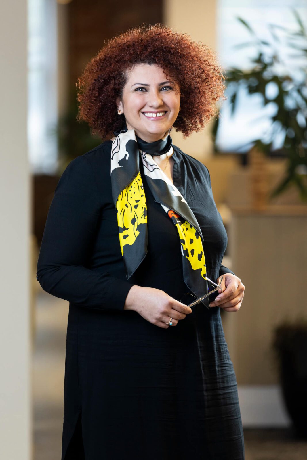 Woman in black dress and black, yellow and white print scarf with reddish brown afro stands holding glasses, smiles broadly to camera