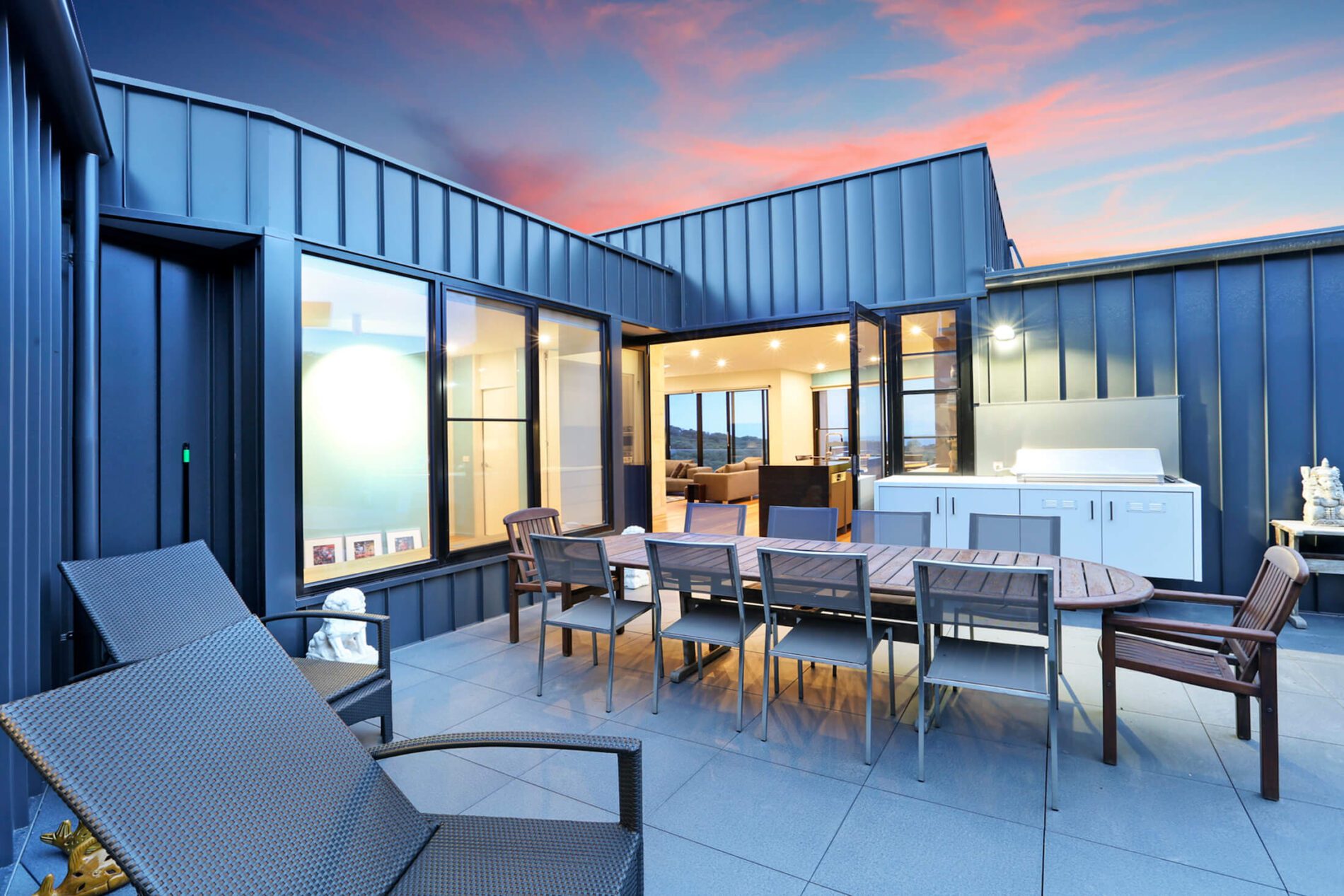 Patio at sunset with standing seam metal building cladding, timber dining table and chairs, open doors to kitchen and living