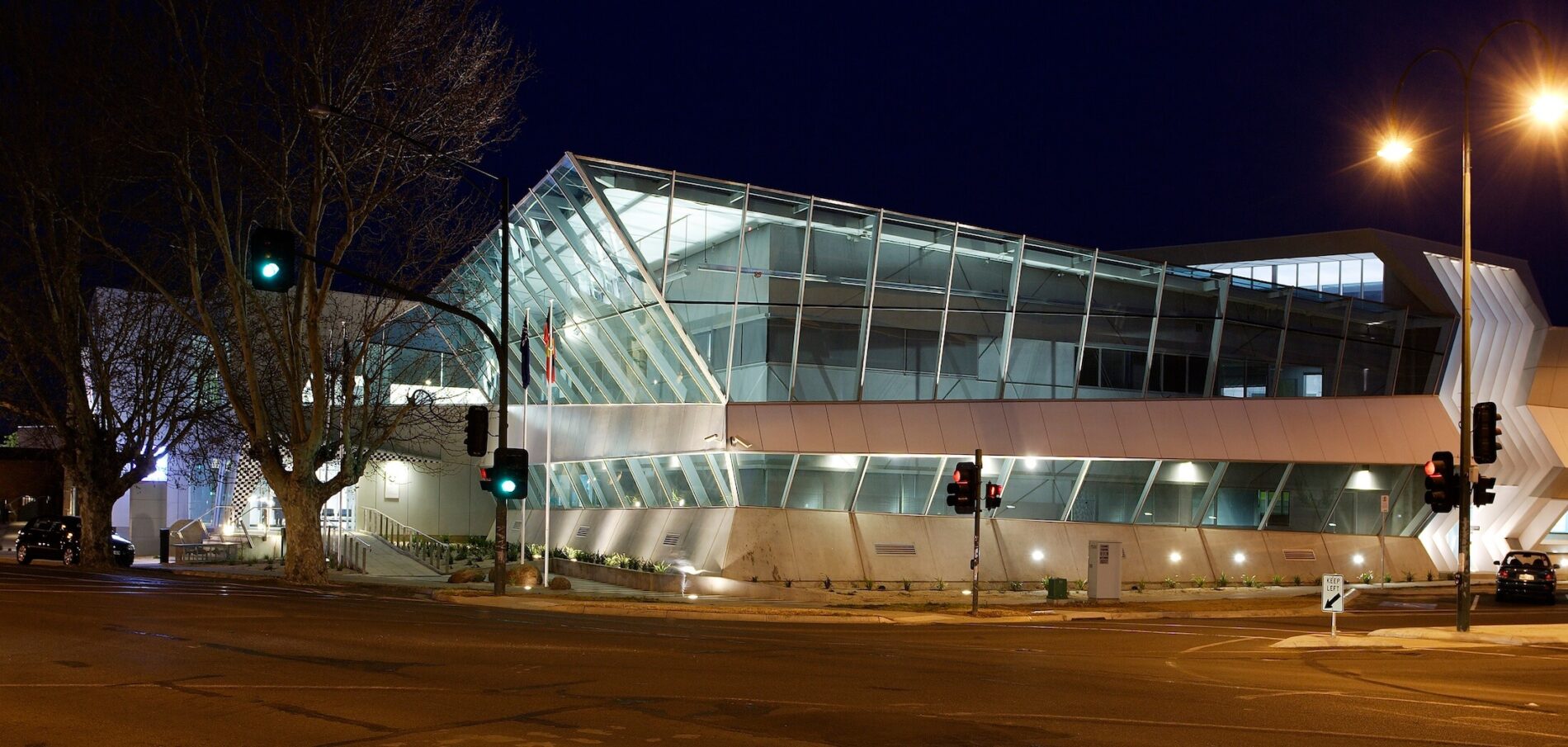 Corner view of angled glazed double facade on large police complex, tree to the left, pictured at night