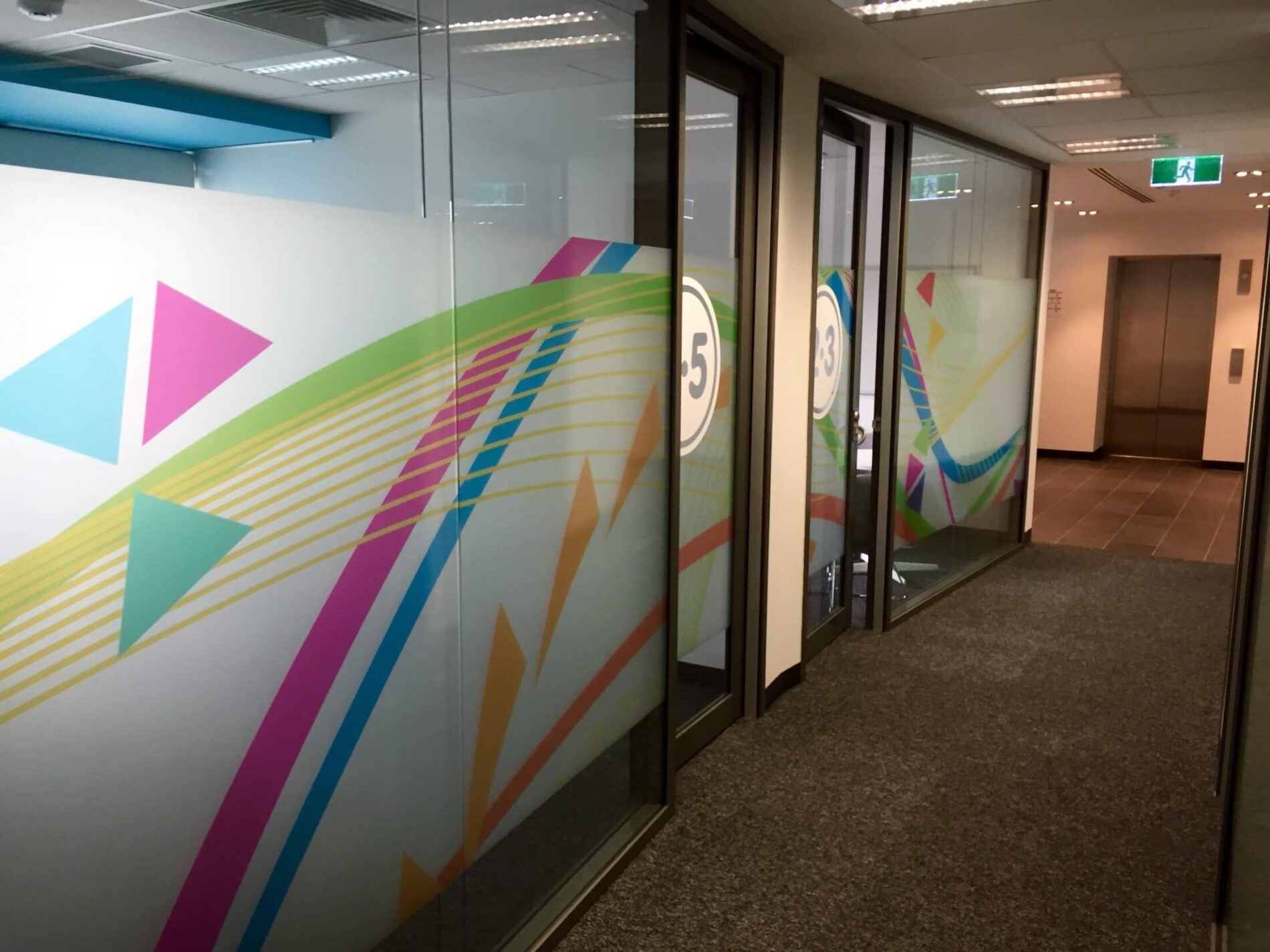 Colourful privacy decals on office glazing alongside hallway leading to lift