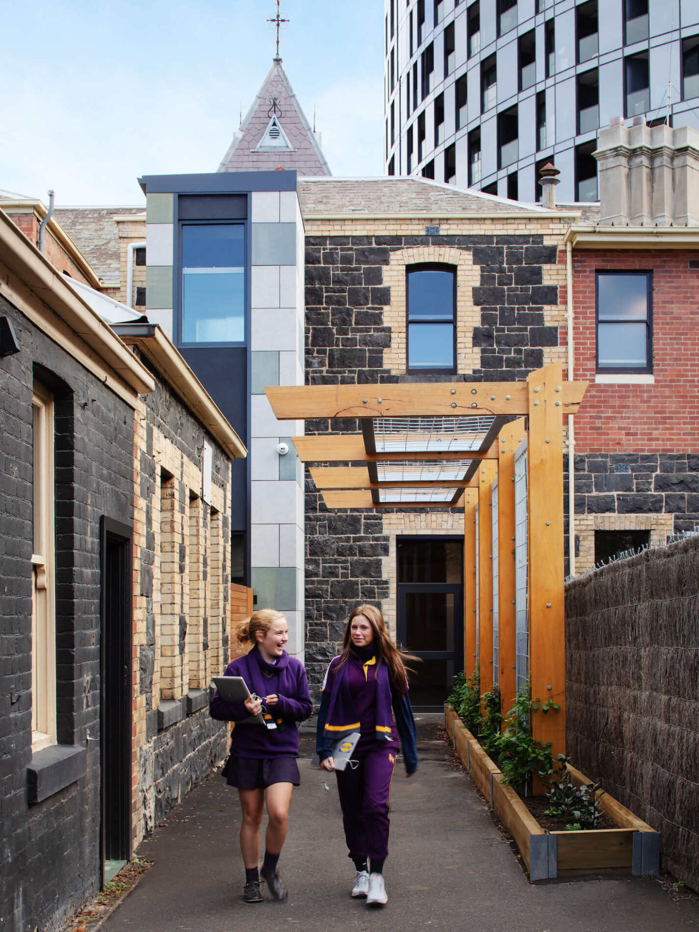 Two students in purple uniforms walk together along outdoor walkway leaving historic bluestone building, timber trellis alongside