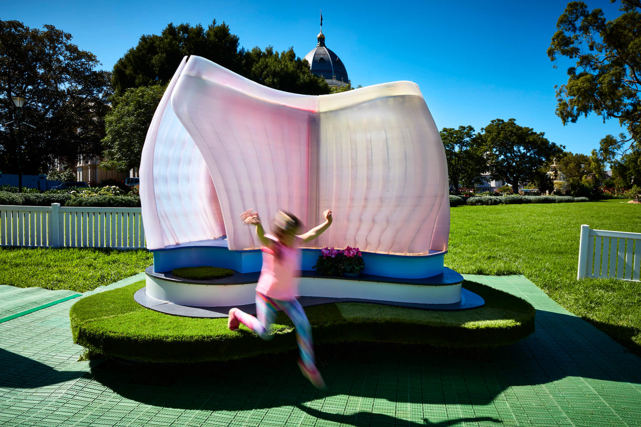 Child leaps from plinth in front of organic pink formed structure with blue sky and trees beyond