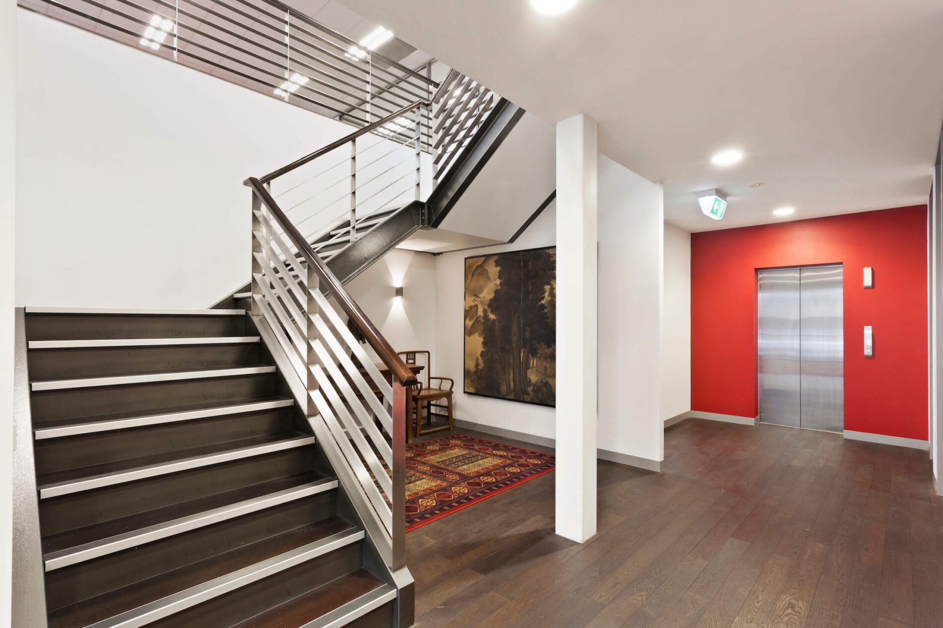 Stairs leading from foyer with timber look flooring, rug-covered stairwell and lift with red feature wall