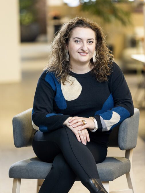 Woman in black and navy dress, with brown curly hair sitting on a blue chair