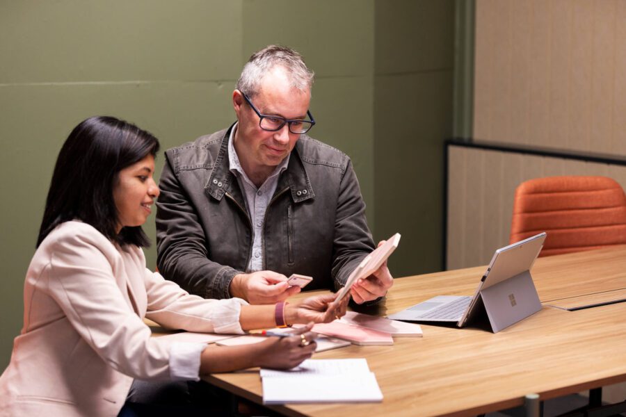 Two architects sit at a table looking at samples in an office with a green wall behind.