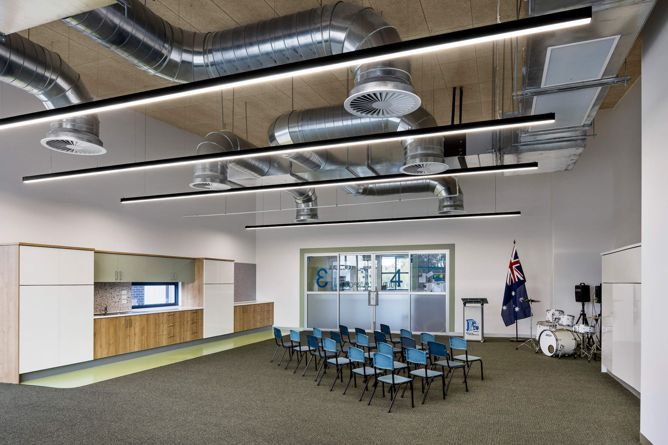 Carpeted classroom with kitchenette, drums and Australian flag, chairs assembled, ducted ventilation above.