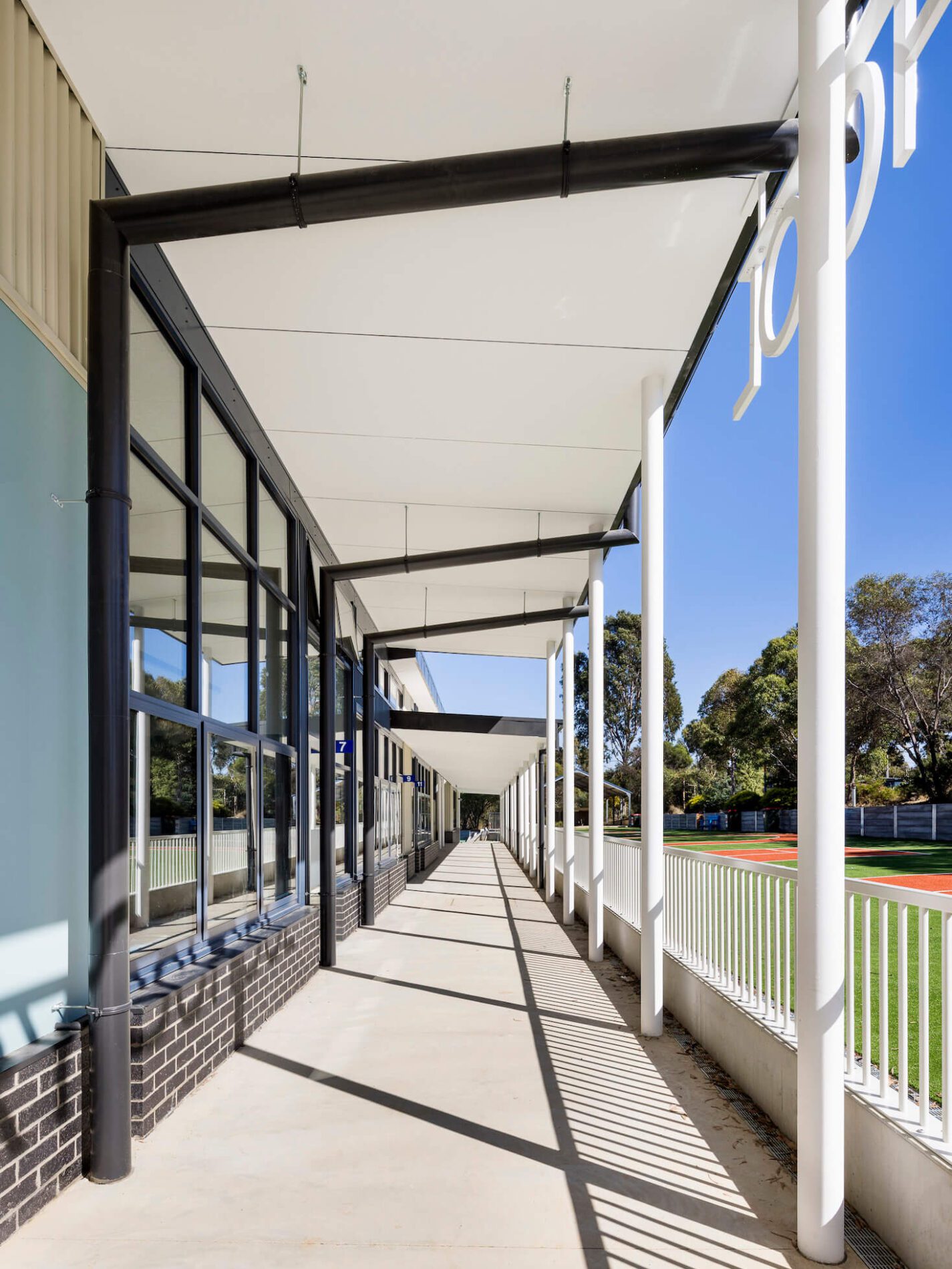 Covered outdoor school walkway with white poles and black trim windows, next to turfed area.