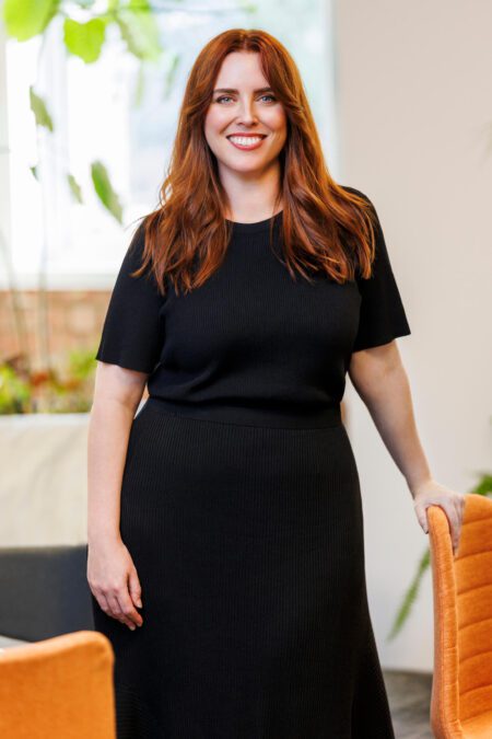 Woman in black outfit with long red hair stands smiling broadly at camera, holding onto orange chair, office plants in background