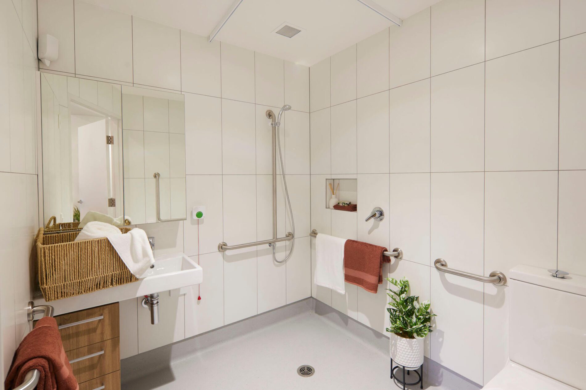 Large format white tiled bathroom with accessible shower and handrails, timber veneer drawers and linen basket