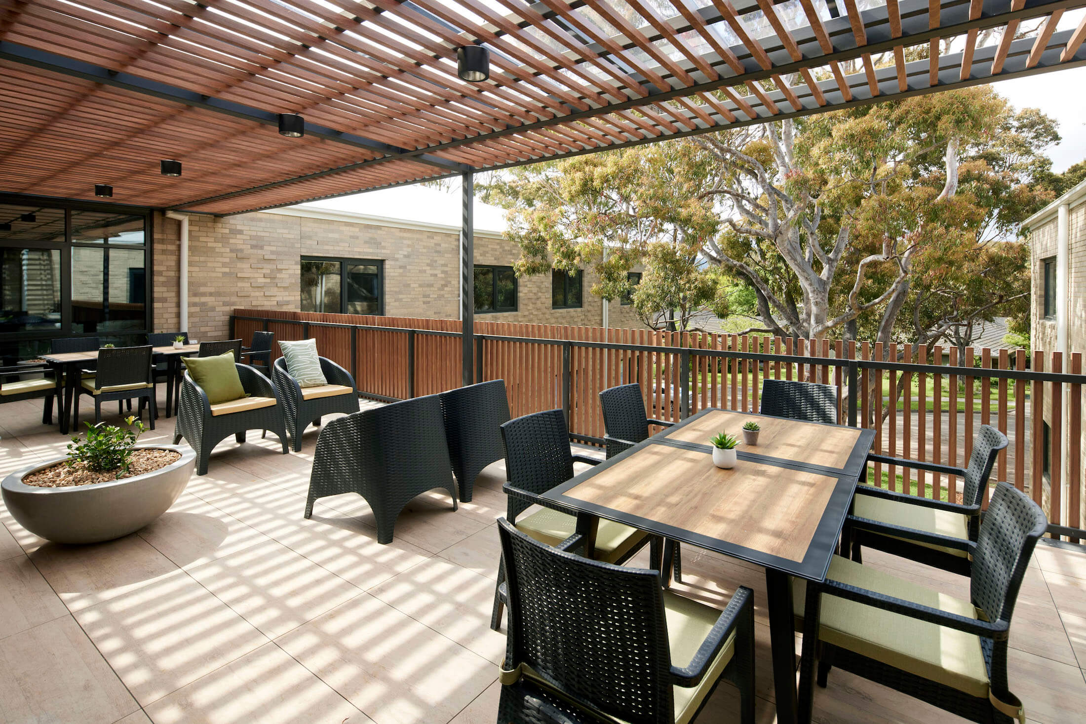 Raised outdoor patio with slatted timber shade cover throwing dappled shade on dining and lounge settings, large gum tree beyond