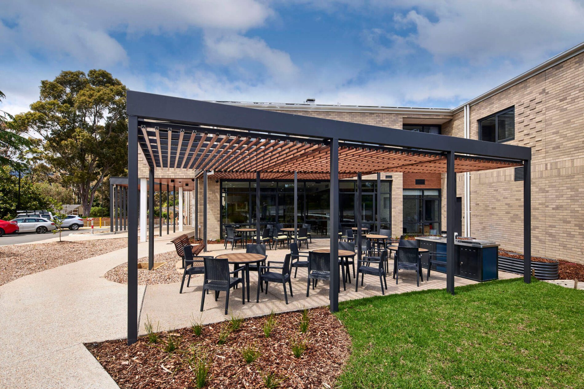 Outdoor cafe style dining and barbecue under a pergola structure, cream brick building in background