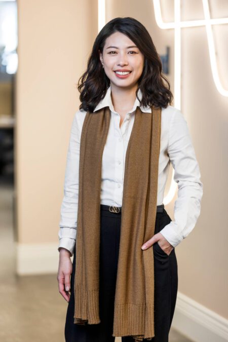 Woman with dark hair, white shirt, brown scarf, stands smiling broadly at camera in front of neon tubing business signage wall