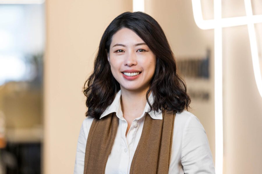 Woman with dark hair, white shirt, brown scarf, stands smiling broadly at camera in front of neon tubing business signage wall