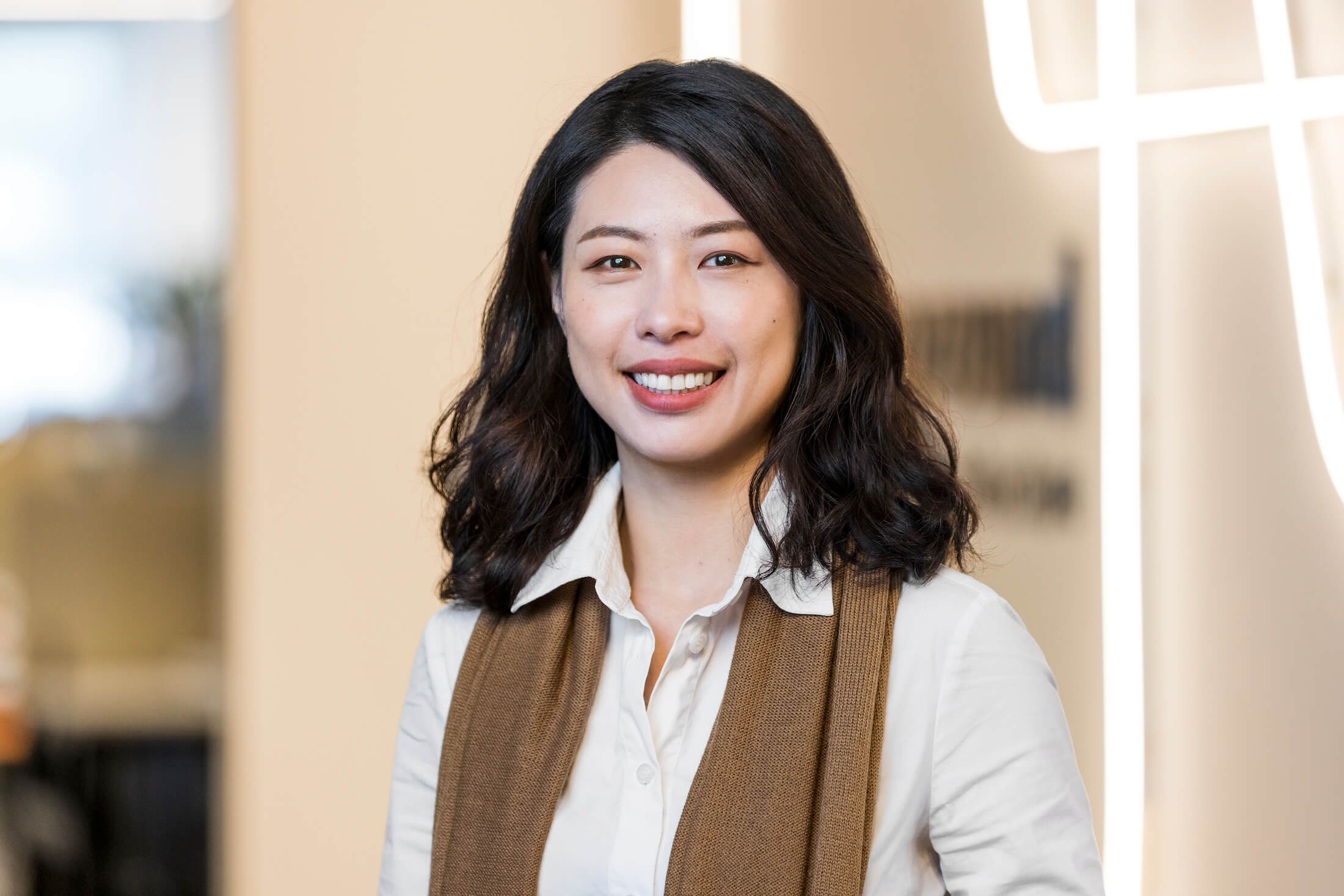 Woman with dark hair, white shirt, brown scarf, stands smiling broadly at camera in front of neon tubing business signage wall