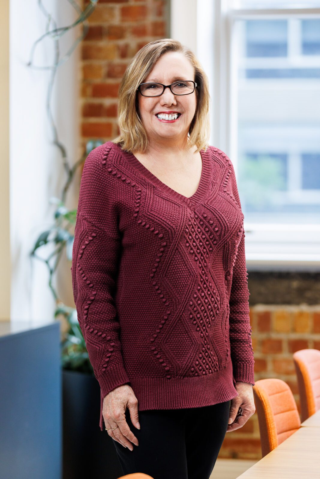 Woman with plum coloured knitted top and glasses smiles at camera