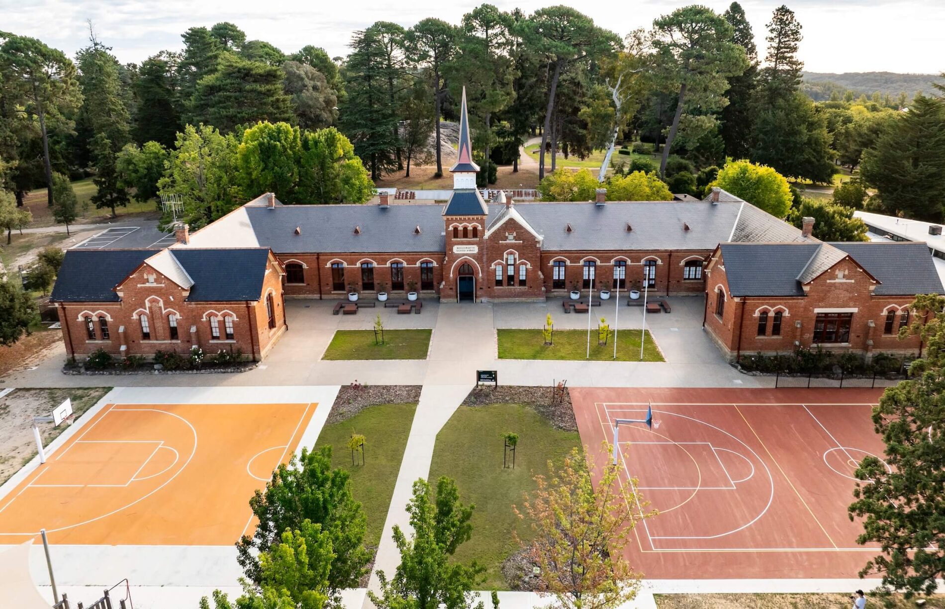 Aerial view of horseshoe shaped heritage brick school building with central spire, grassed and sports surfaced forecourt