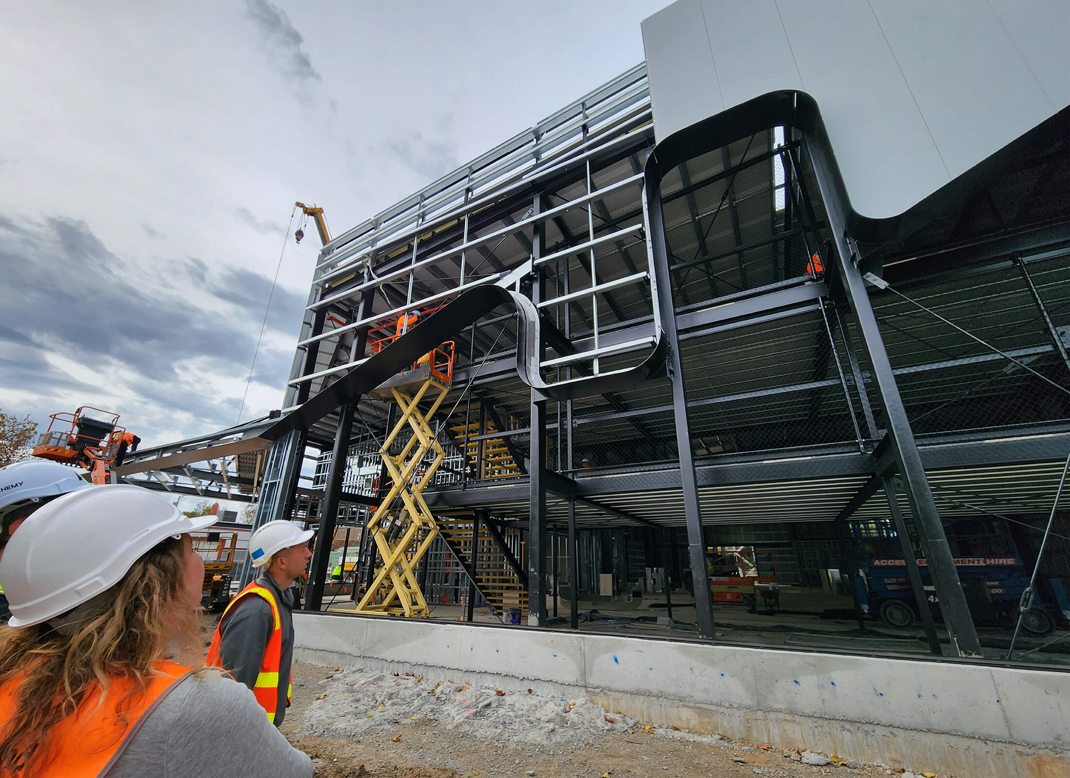 Construction progress shot from site, showing black steel framing. People with white hard hats and yellow high vis vests stand in the foreground.