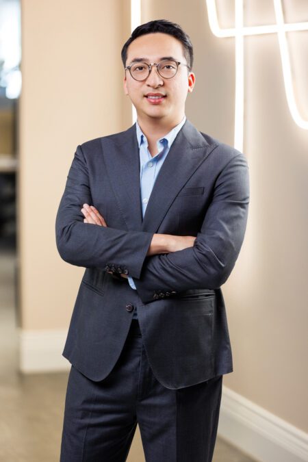 Man wearing glasses and a suit standing in an office with his arms crossed